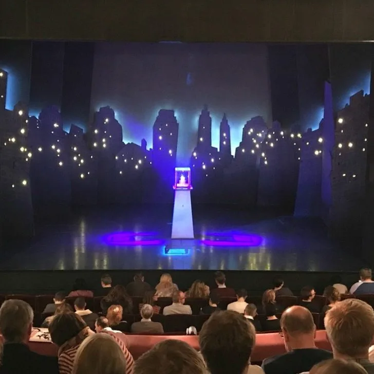 A group is sitting in a room with a stage and a large screen.