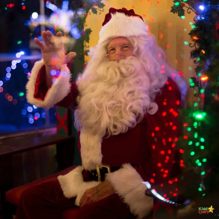 Santa Claus is spreading holiday cheer in a festive indoor setting, surrounded by a Christmas tree adorned with colorful decorations, lights, and ornaments.