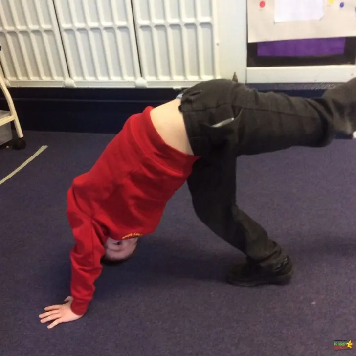 A person is balancing on one knee with their elbow and hip joints in the air, wearing red active pants and a yoga pant, demonstrating physical fitness indoors on a Kiddy Charte.