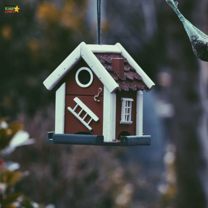 A birdhouse hangs from a tree in front of a winter-covered house, providing a cozy outdoor home for birds in the cold season.