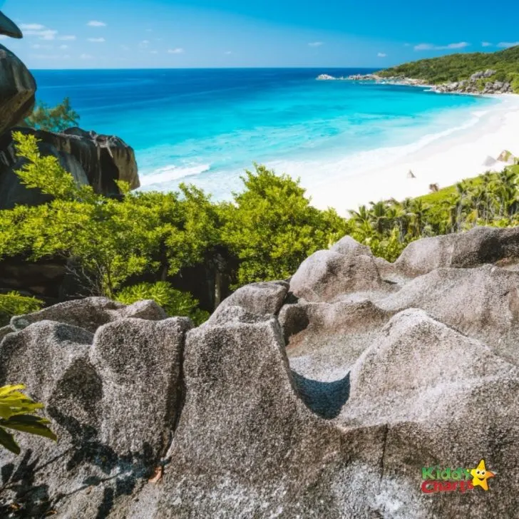 A lush tropical landscape of azure ocean, rocky coast, and lush green trees frames a picturesque Caribbean bay, dotted with clouds and islands in the distance.