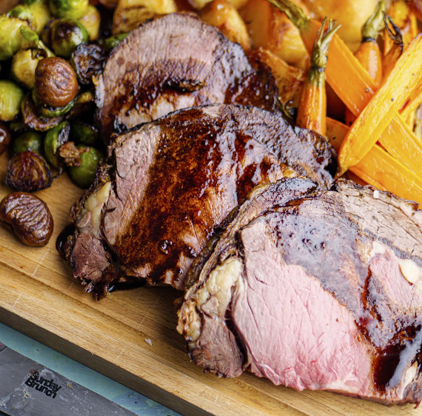 The chef is roasting a variety of meats, including duck and turkey, and carving them with animal fat while surrounded by a spread of produce and hendl for a Thanksgiving feast.