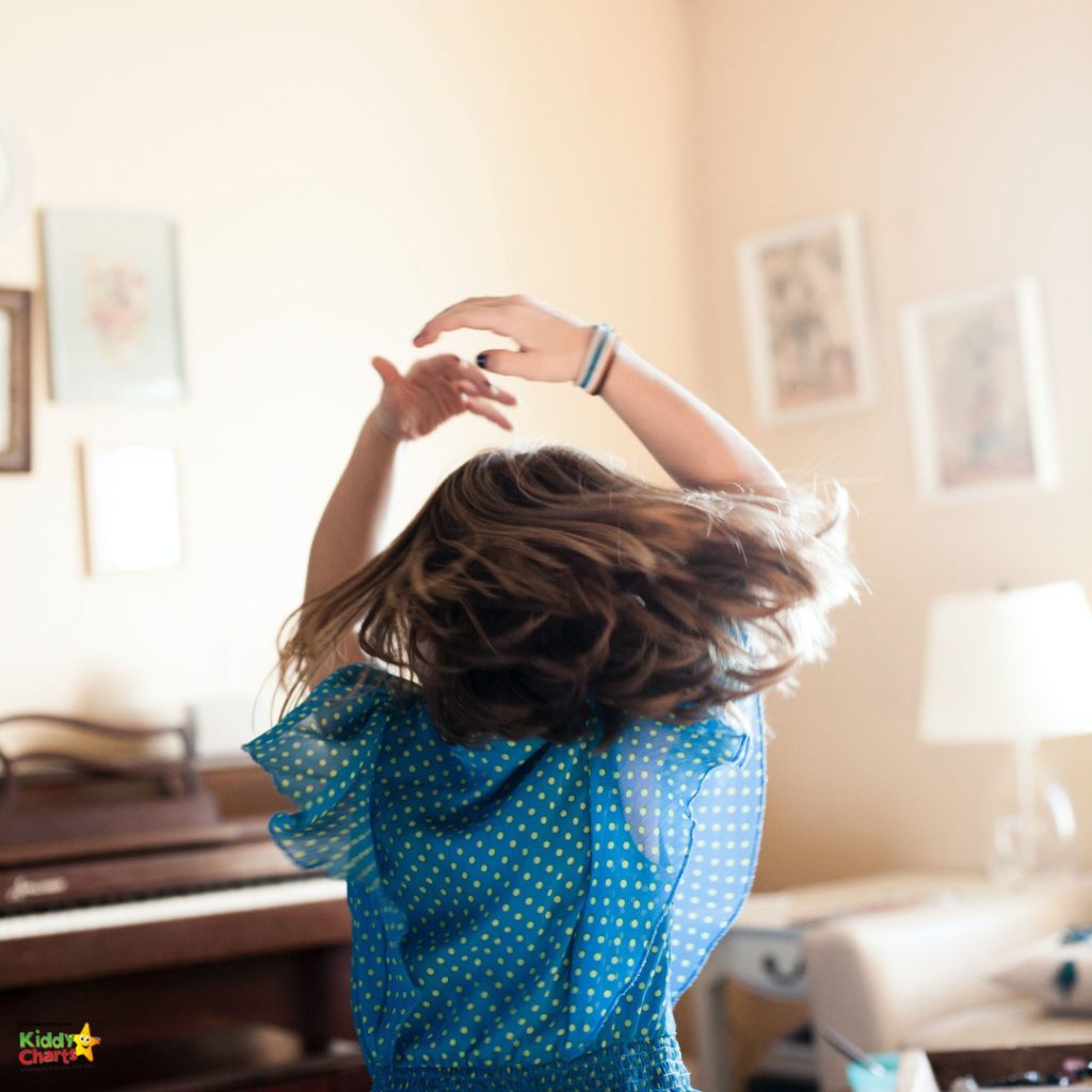 A child in a blue polka-dotted dress is joyfully twirling, with hair flying, in a warmly lit room featuring elegant furniture and framed artwork.