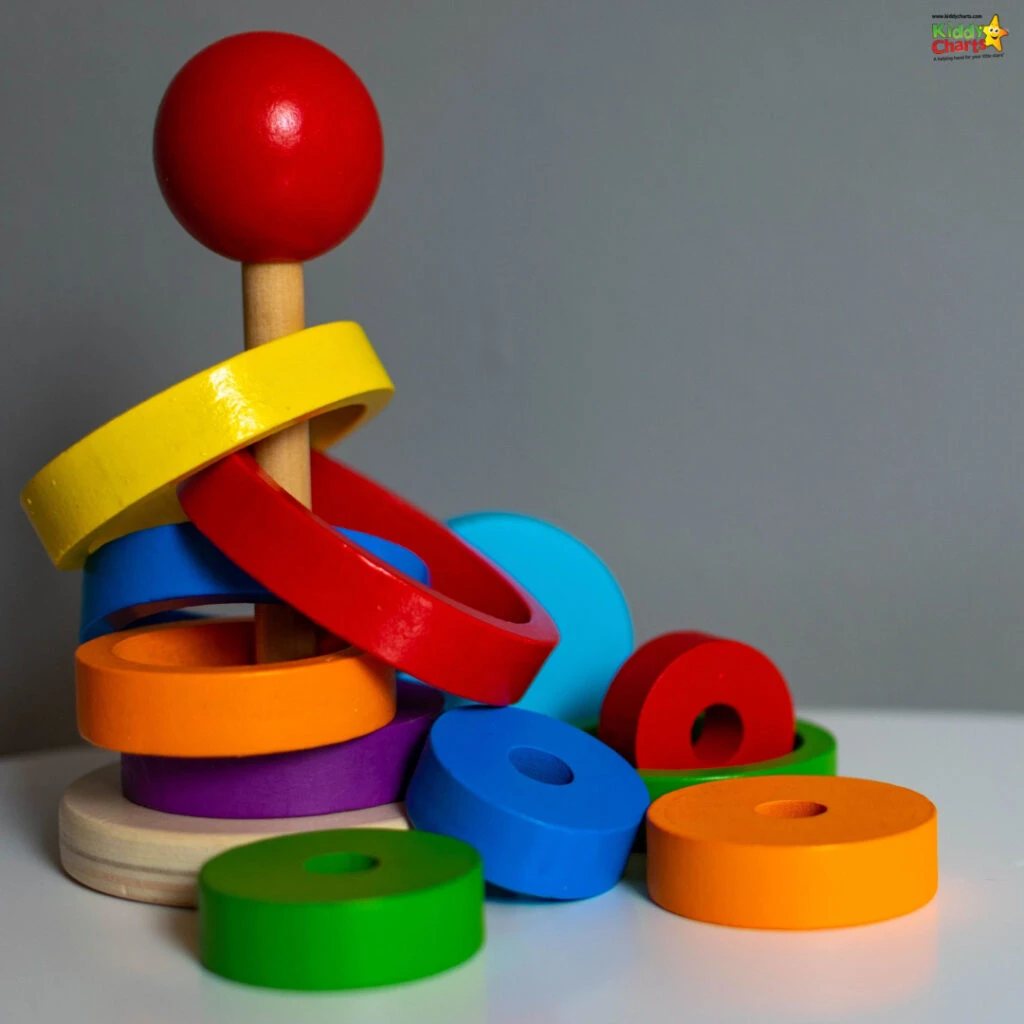 Colorful wooden rings and shapes are stacked haphazardly on a pole against a grey background, creating a vibrant display reminiscent of a child's toy.