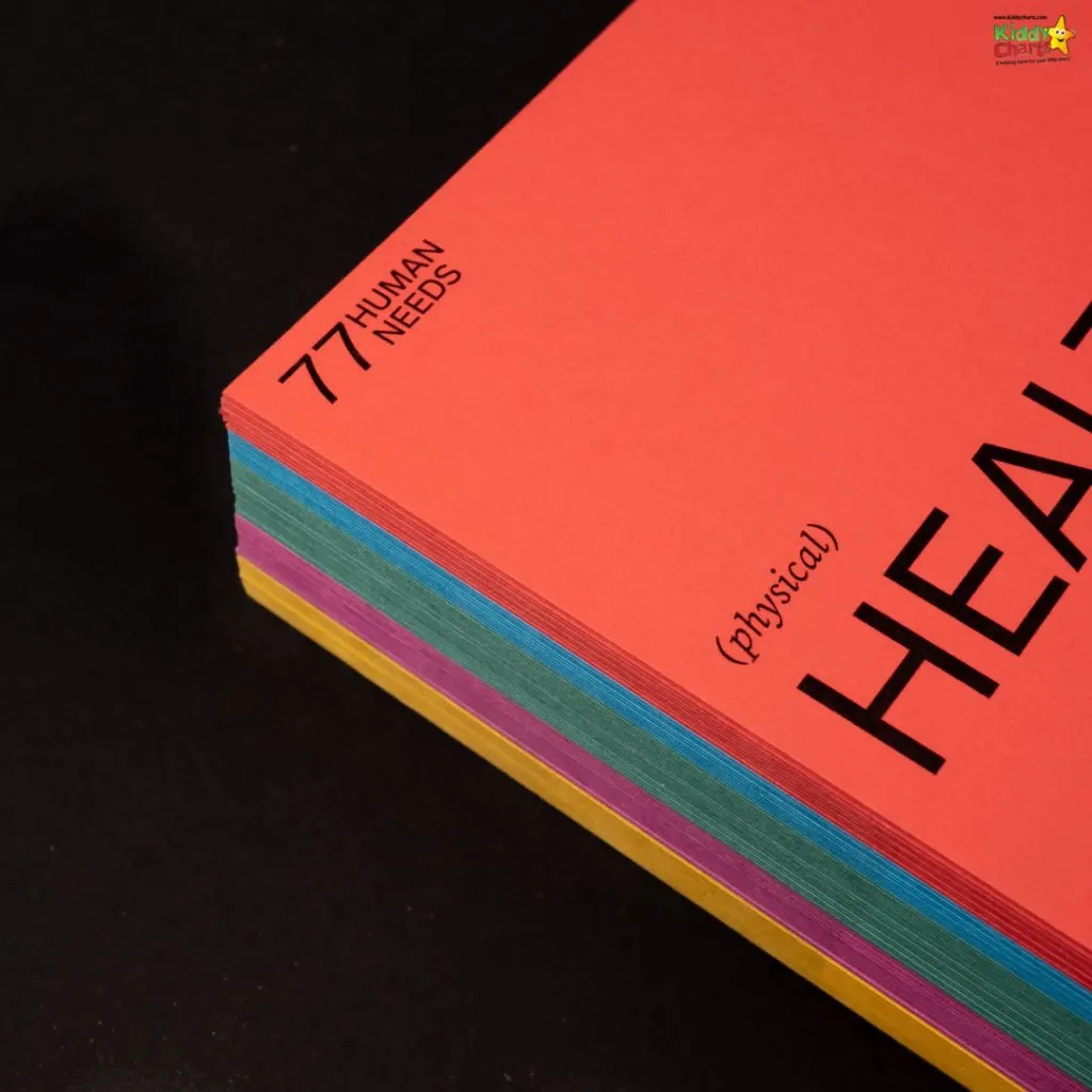 A red book titled "77 Human Needs (physical)" with multicolored page edges is placed on a black surface, angled to show the spine.