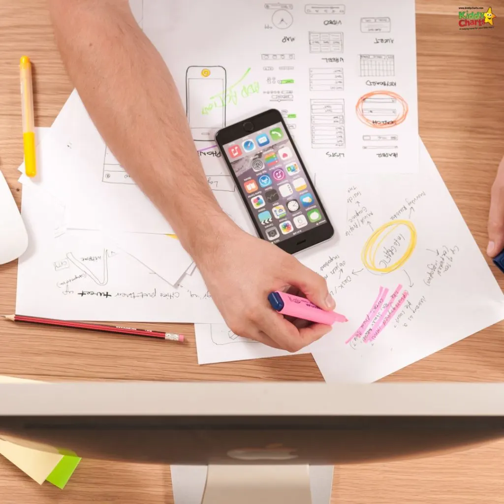 A person is working on app design planning, surrounded by sketches and notes, highlighting with a pink marker, with a smartphone and monitor visible.