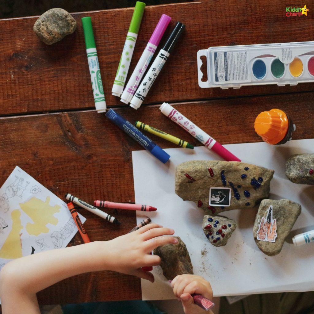 This image shows a child's hands decorating rocks with markers and gems on a wooden surface surrounded by crayons, painted rocks, and a watercolor palette.