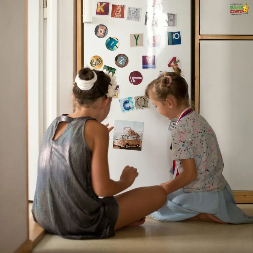 Two children are sitting on a wooden floor, engaging with a magnetic board that has a variety of colorful letter magnets and a picture.