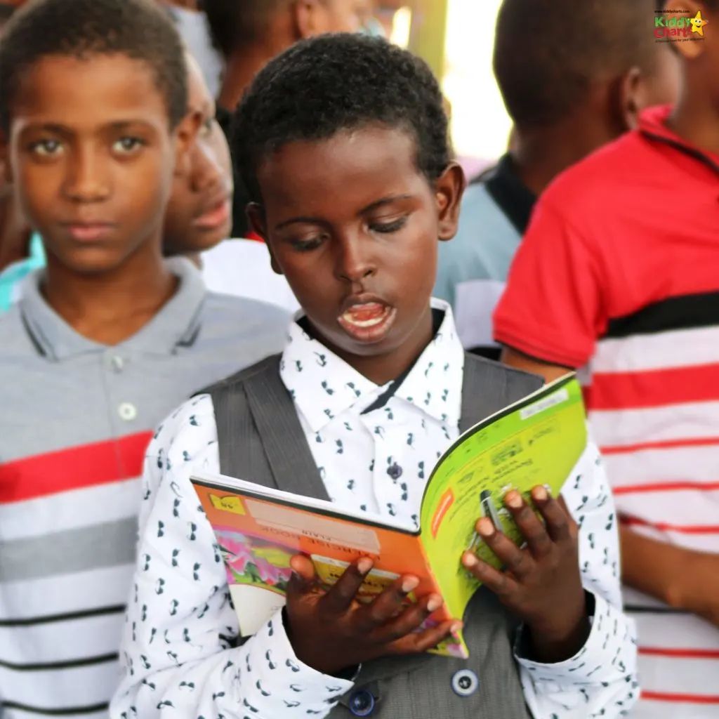 A child in a patterned shirt is focused on reading a book, among other children in a group, seemingly in an informal gathering.