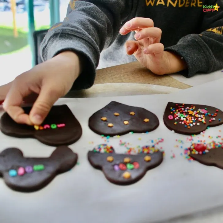 A person is decorating heart-shaped chocolate cookies with colorful sprinkles on parchment paper, creating a fun, homemade baking scene.