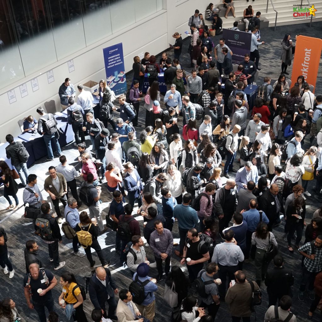 The image shows a crowded indoor event with numerous individuals standing and conversing, some holding beverages, and banners visible, indicating a professional or corporate gathering.