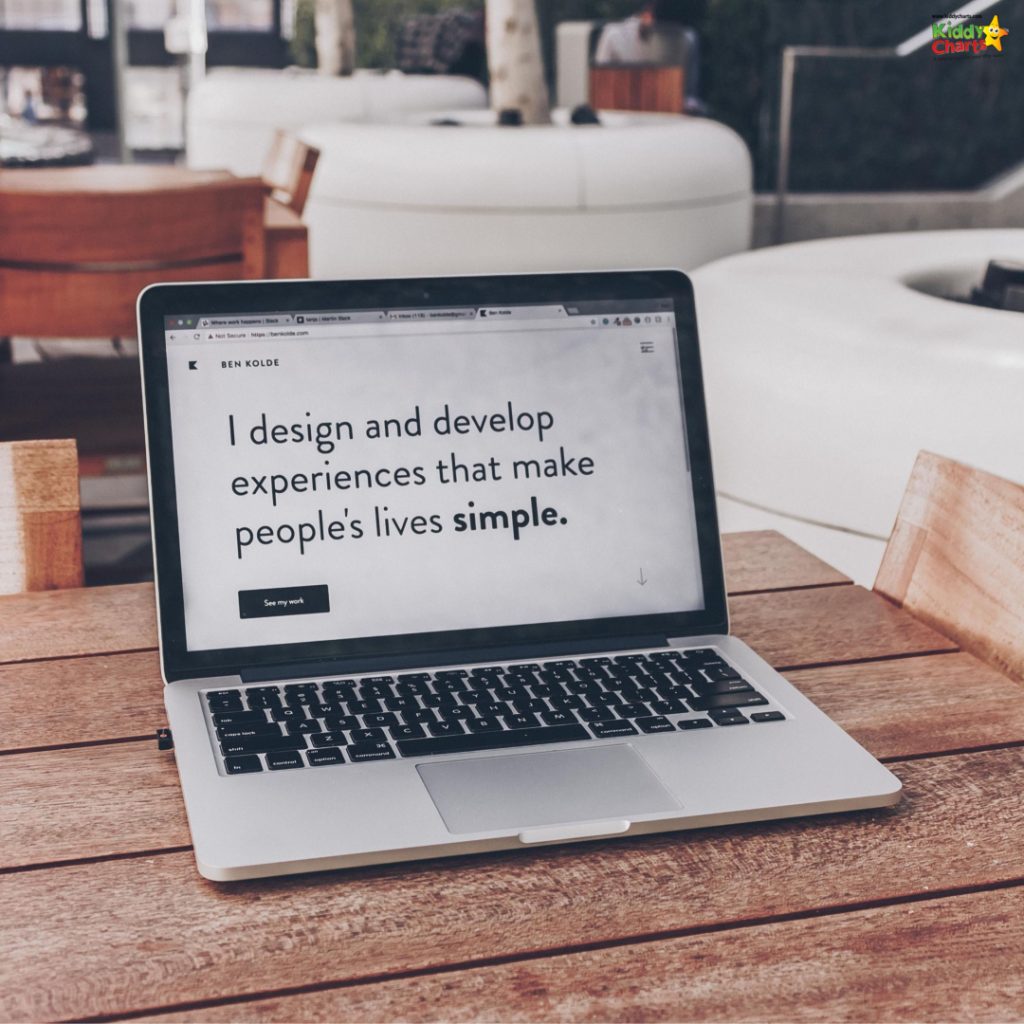 An open laptop on a wooden table displays a website with text about designing and developing simple experiences for people, outdoors with soft-focused background.