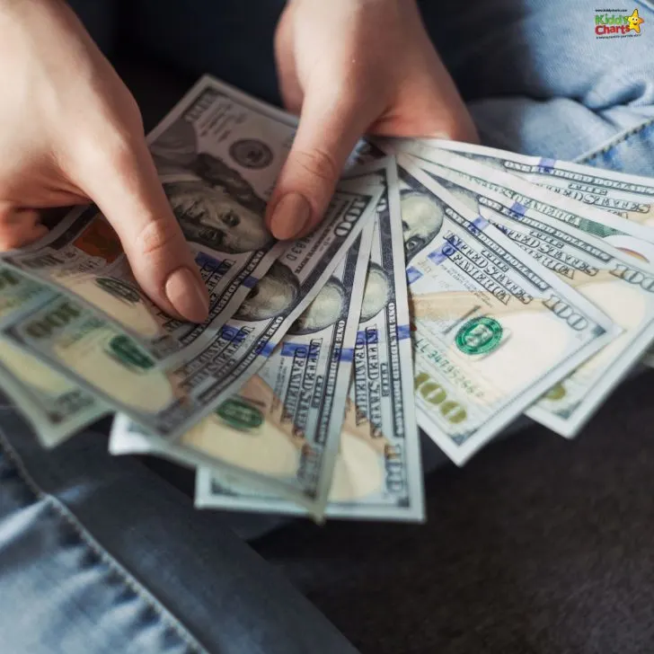 A person is holding several U.S. 100-dollar bills fanned out in their hands, with denim clothing partially visible in the background.