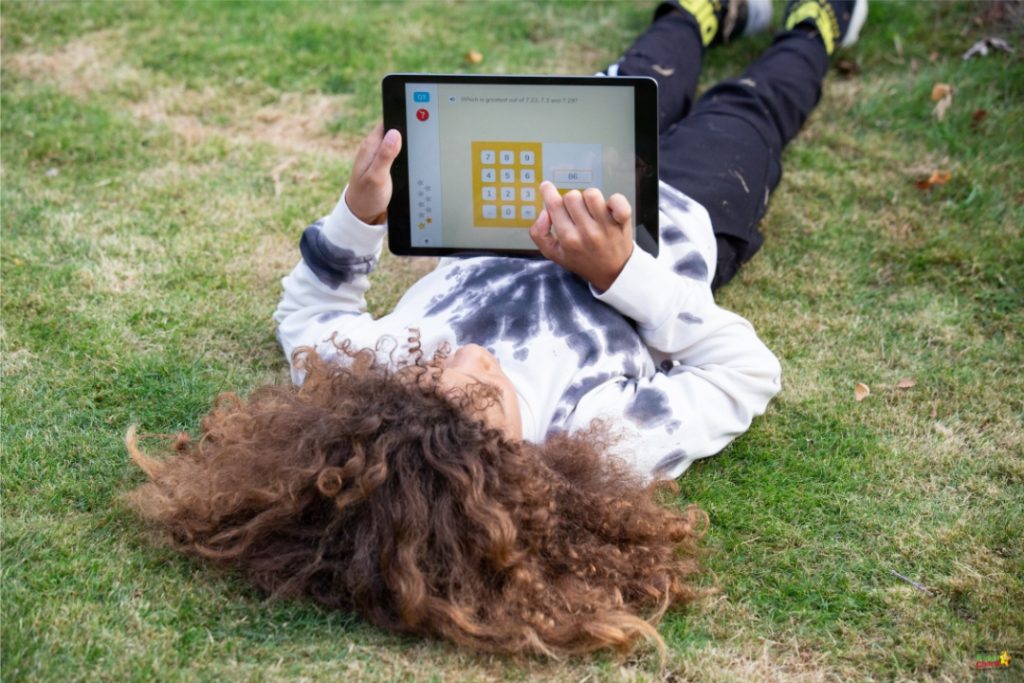 A person lies on grass, holding a tablet with a math problem on the screen, wearing a tie-dye sweatshirt and black pants with sneakers.