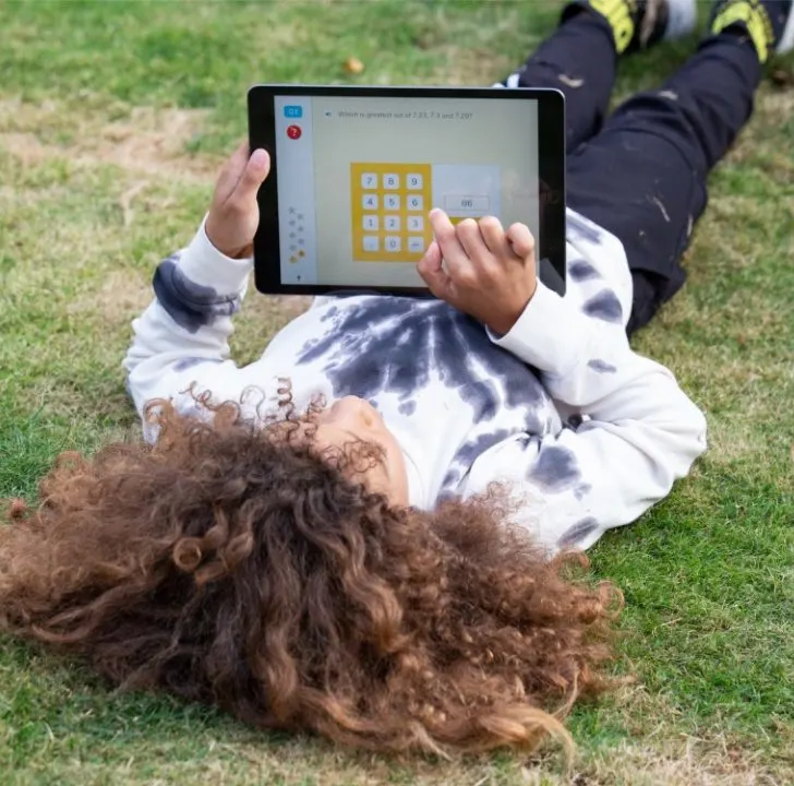 A person lies on grass, holding a tablet with a math problem on the screen, wearing a tie-dye sweatshirt and black pants with sneakers.