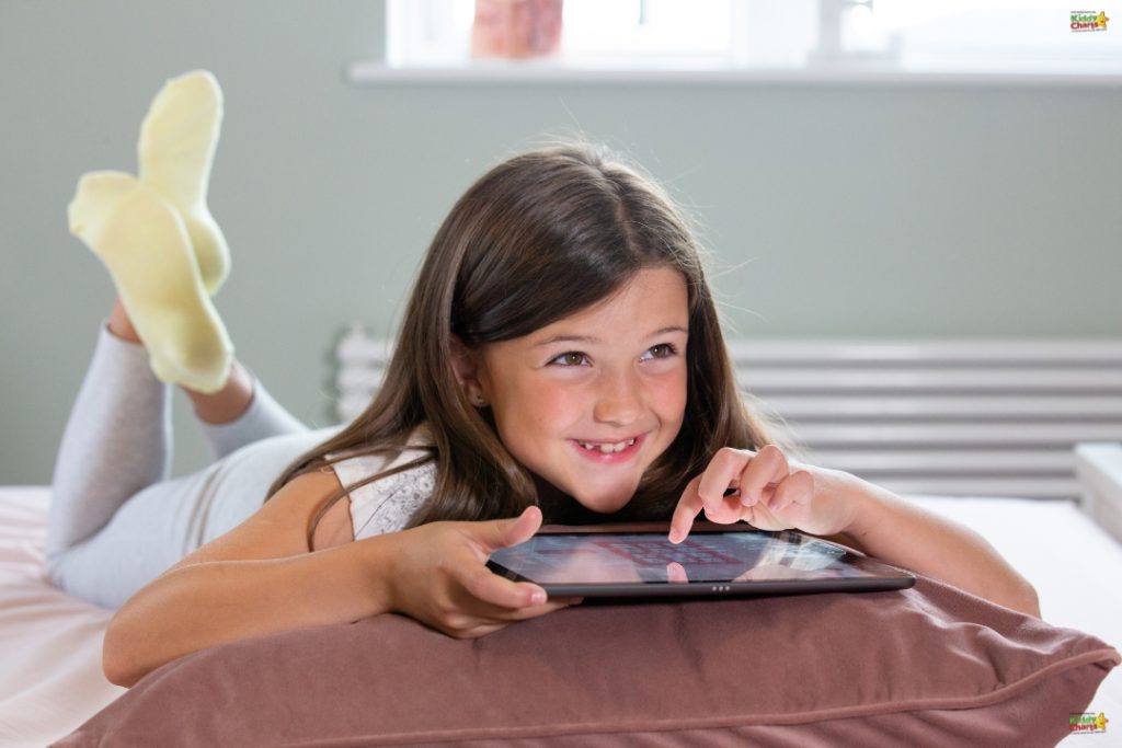 A young girl is lying on her stomach on a bed, smiling while using a tablet, wearing yellow socks and casual clothing in a bright room.